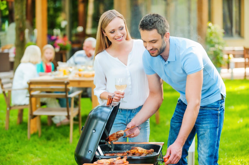 Family barbeque. Happy young couple barbecuing meat on the grill while other members of family sitting at the dining table in the background